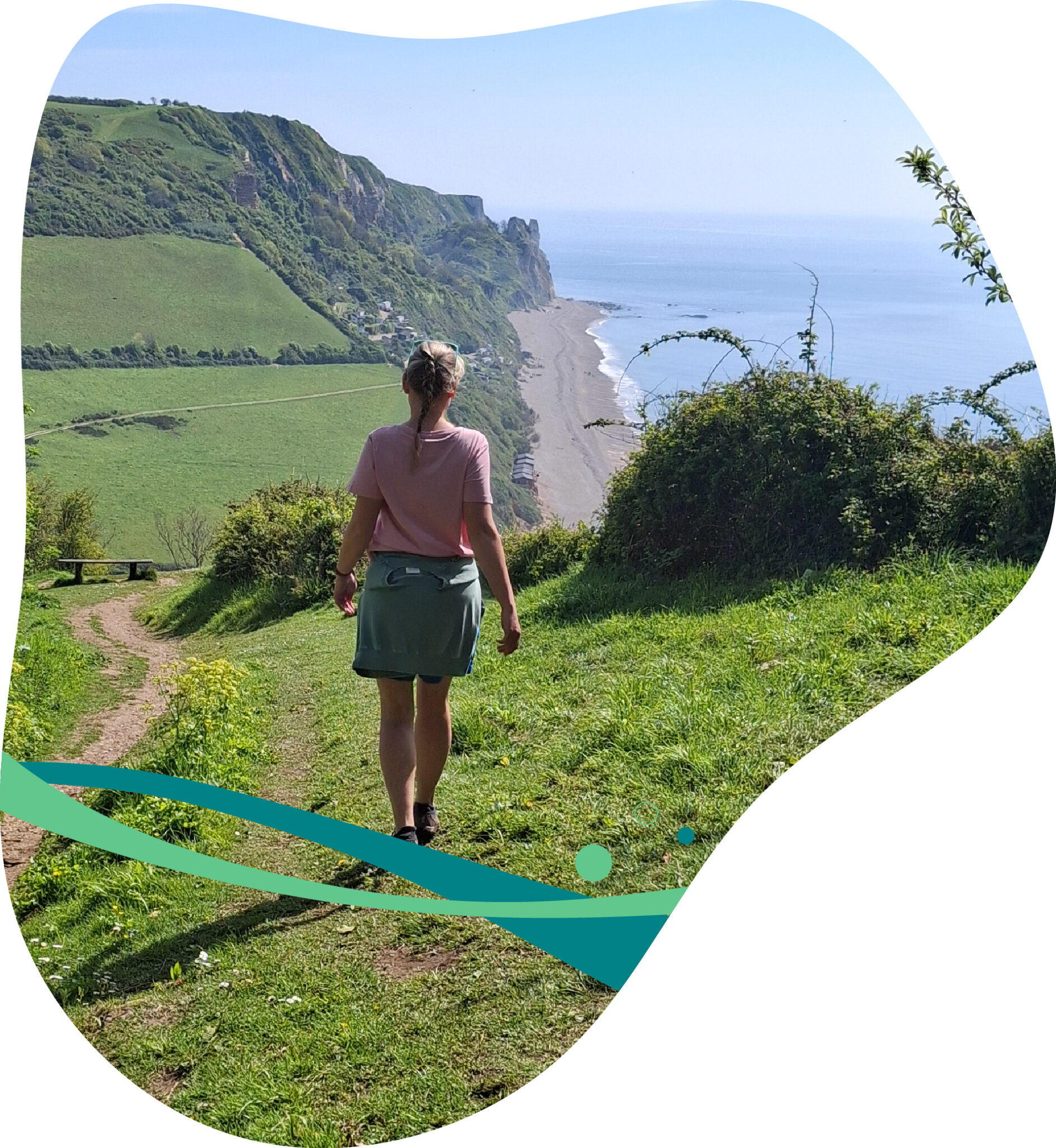 A woman walks along a grassy coastal path overlooking cliffs and the sea on a clear sunny day