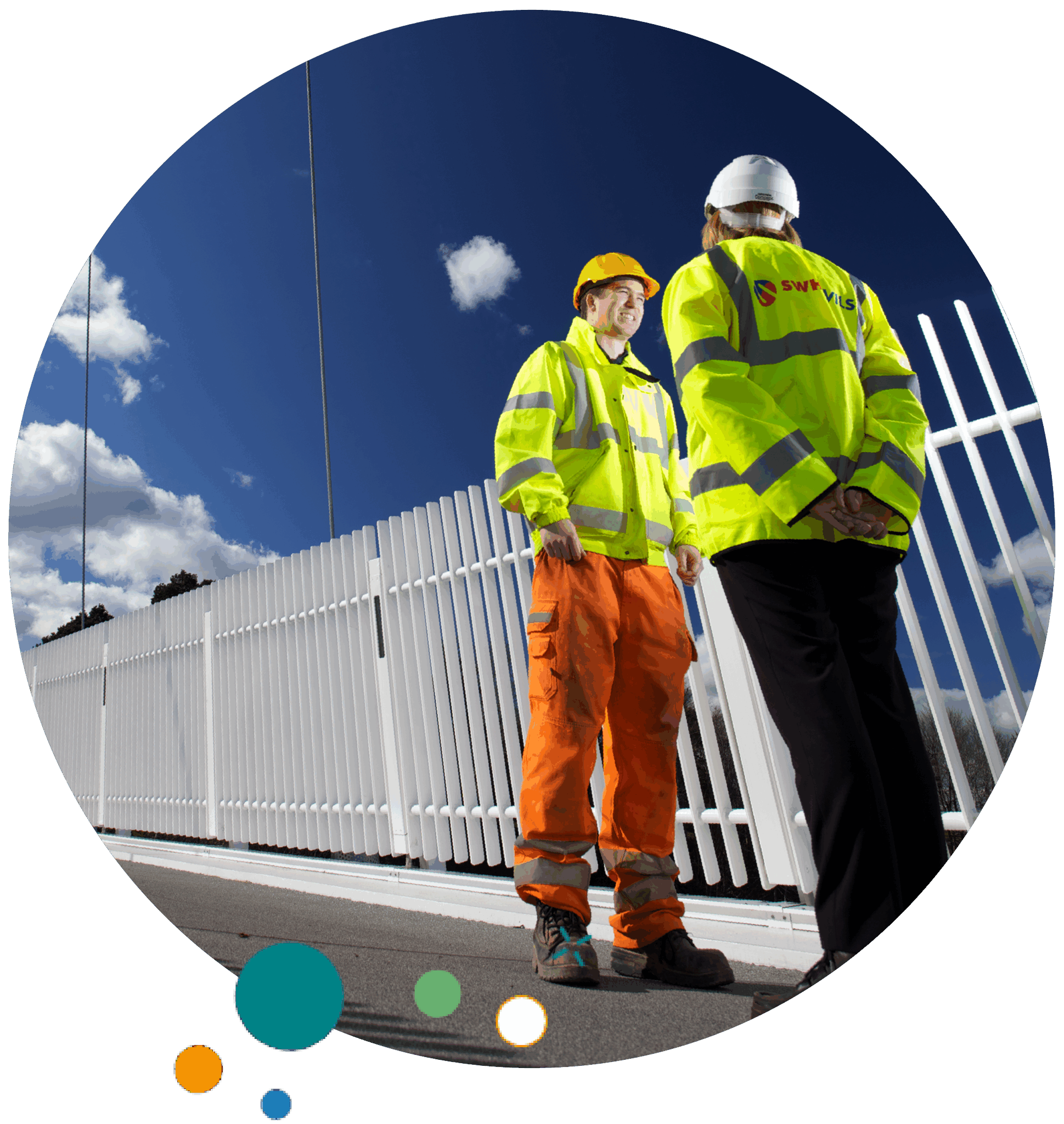Two construction workers in high-visibility jackets and helmets stand talking outdoors beside a white fence under a bright blue sky with clouds.