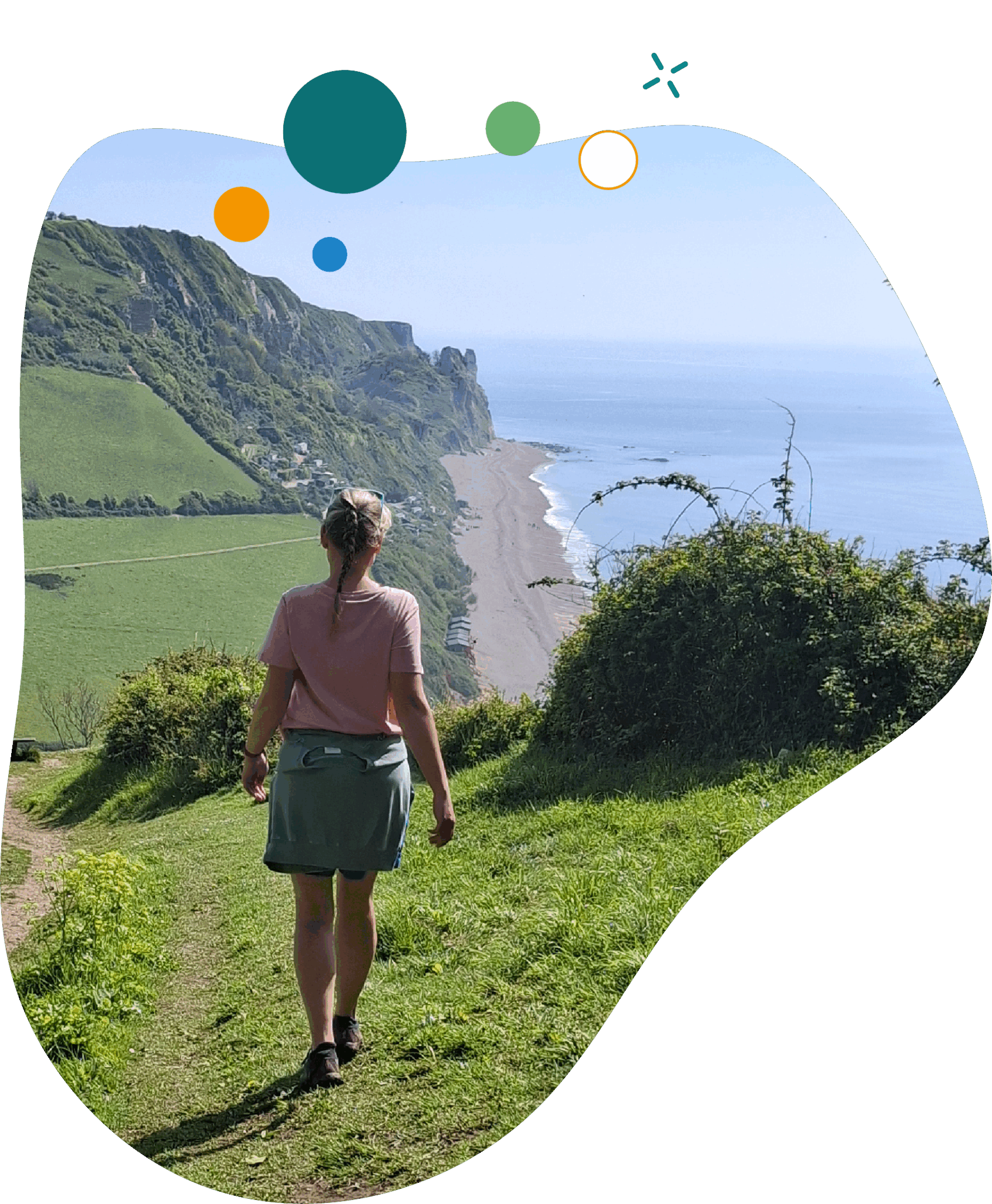 A person with braided hair, wearing a pink shirt and a light jacket tied around their waist, walks along a grassy cliffside path overlooking the sea. The coastline stretches into the distance with green hills on the left and a sandy beach below. The sky is clear and blue, suggesting a sunny day.