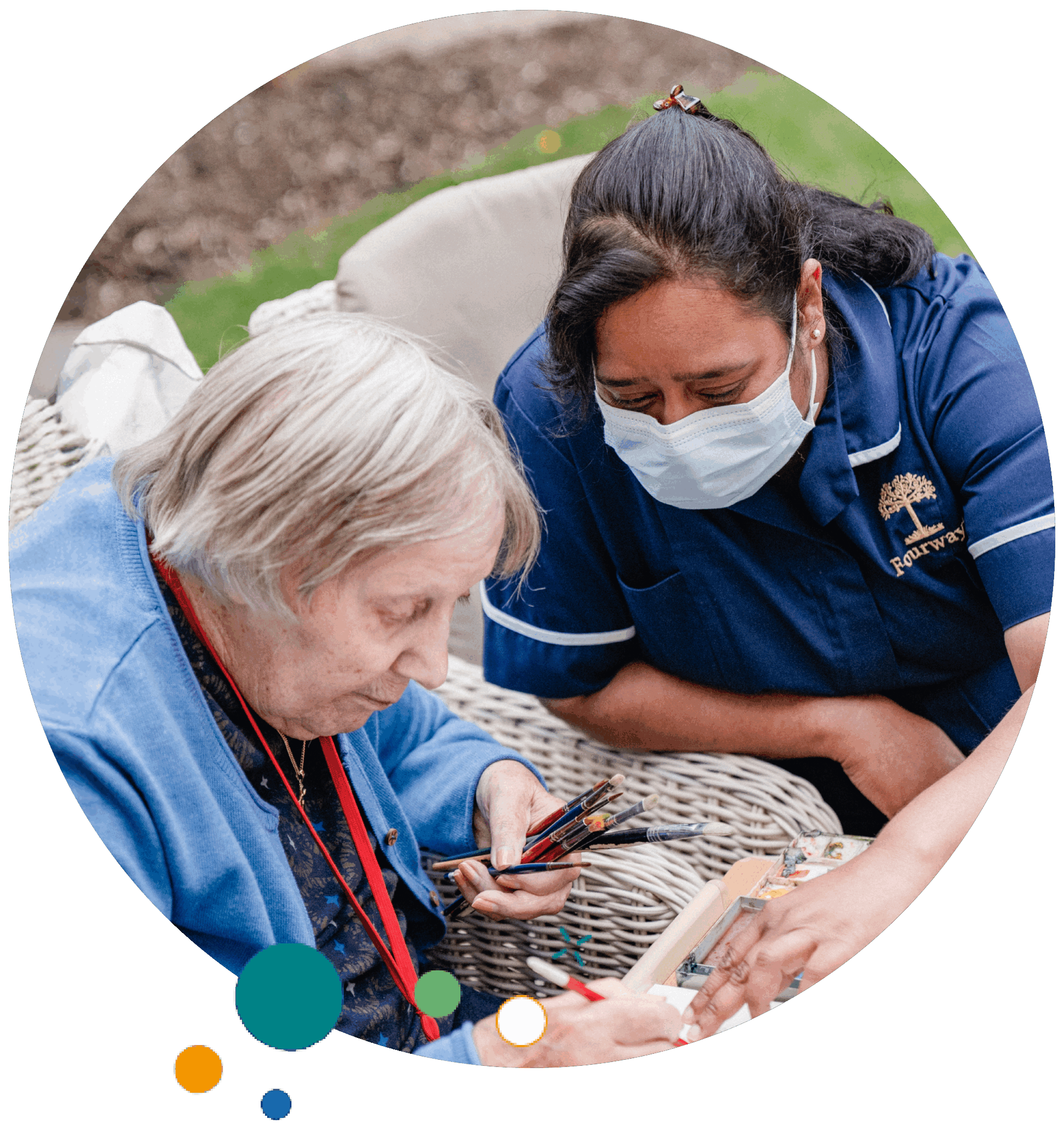A care worker in a blue uniform and face mask supports an elderly woman holding paintbrushes while doing an art activity outdoors.