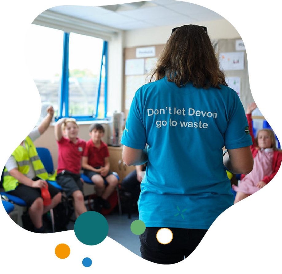 A person wearing a blue shirt with the words 'Don’t let Devon go to waste' speaks to a group of school children sitting in a classroom, some with hands raised to participate