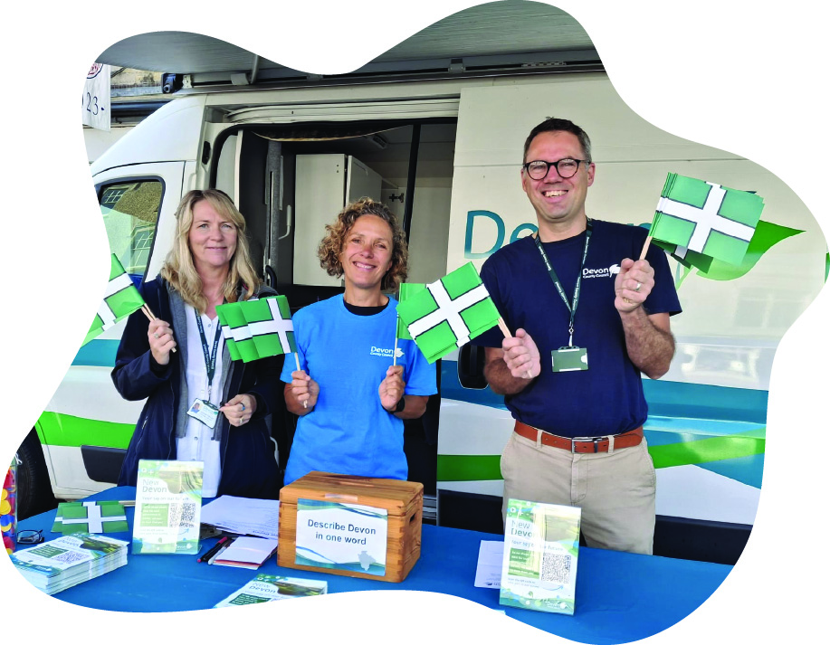 Three people standing behind an information table in front of a branded Devon County Council van, holding green and white flags with a white cross. The table is covered with a blue cloth displaying the word 'Devon' and has brochures, a wooden suggestion box labelled 'Describe Devon in one word,' and a jar filled with colourful tokens.