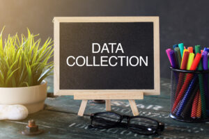 Image of a small blackboard on a desk with the words ' data collection' written in white. On the desk there are also pens in the background part of a plant and a pair of glasses.