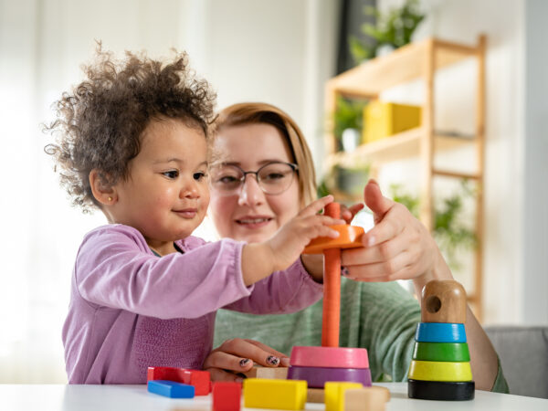 A young woman in a green top is playing with a young child in a pink top stacking rings on a pole.