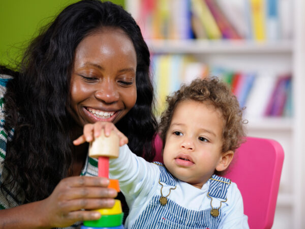 A woman smiles whilst a small child sits on a pink chair playing with a toy. There are books in the background.