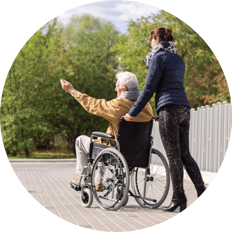 Image is cropped to a circle and shows the backs of two people. One is sat in a wheelchair feeding birds and the other person is pushing the wheelchair.