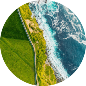 Image is cropped to a circle and shows a birds eye view of the coastline and a green field.