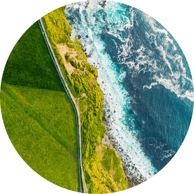 Image is cropped to a circle and shows a birds eye view of the coastline and a green field.