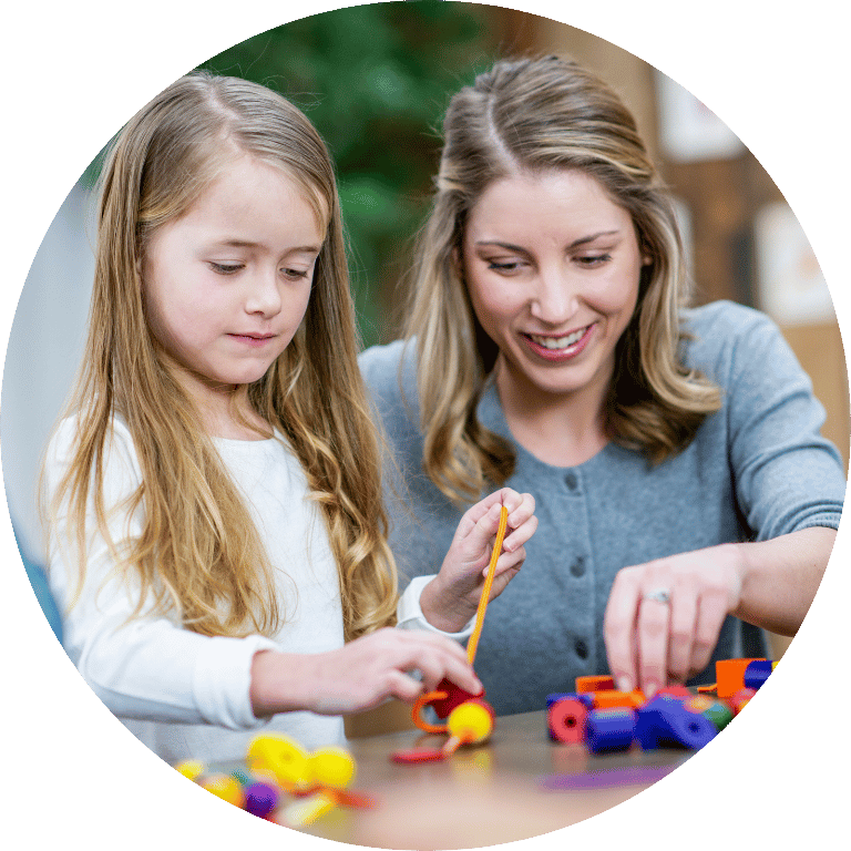 Image is cropped to a circle and shows a young girl playing with toys on a table with an adult.