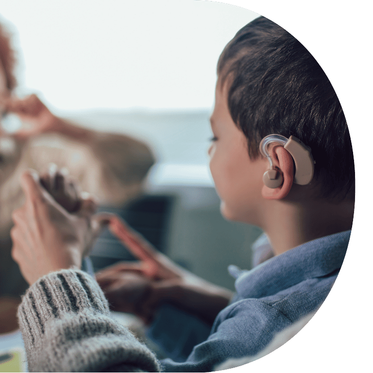 Image is cropped to a D shape and shows the side profile of a child wearing hearing aids and doing sign language.