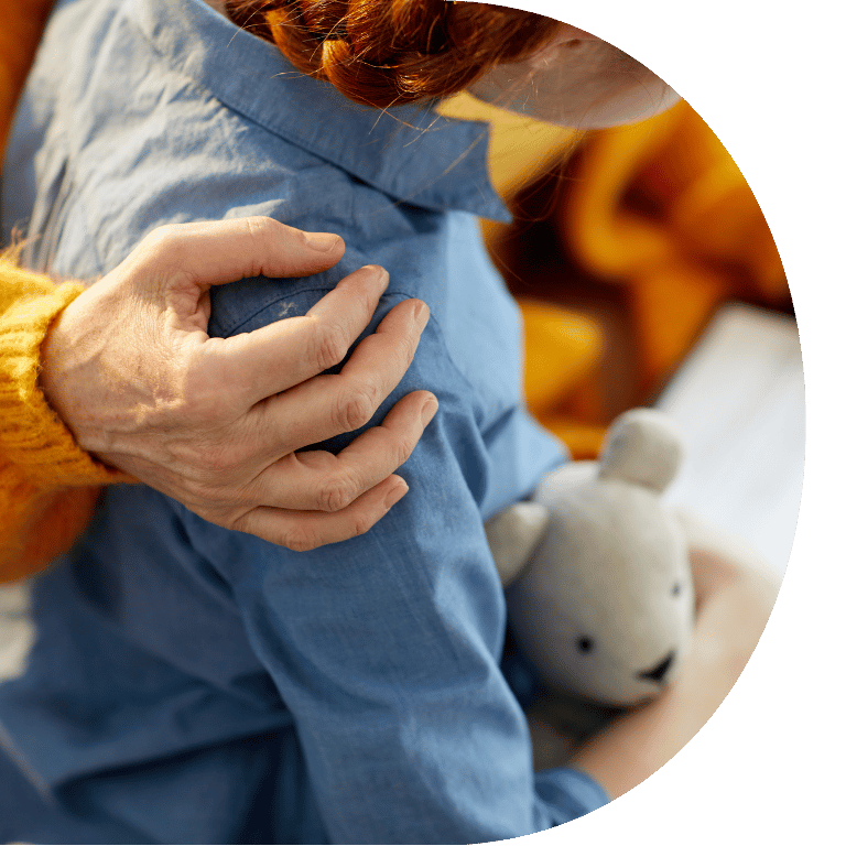 The image is cropped in a D shape and shows a close up of a hand on a child's shoulder