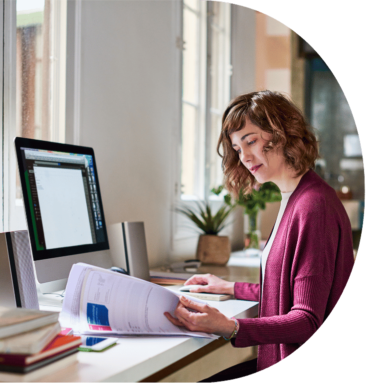 The image is cropped in a D shape and shows a woman looking through paperwork in front of a computer.