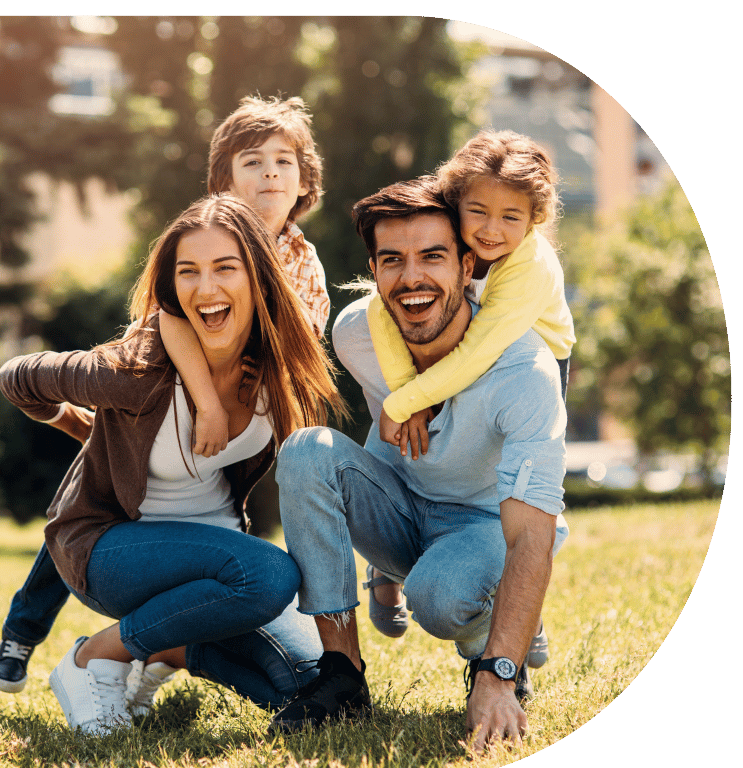 Picture of two adults are crouched down on the grass and two children are hugging them around their necks. The family are smiling at the camera.