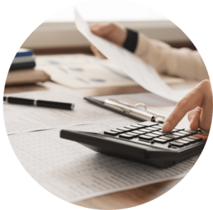 This is a picture of a desk with documents and a calculator on it. Someone is typing on the calculator, holding up one of the documents in the other hand. Only the hands are shown in the shot. The picture is in the shape of the letter O.