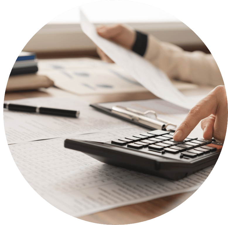 This is a picture of a desk with documents and a calculator on it. Someone is typing on the calculator, holding up one of the documents in the other hand. Only the hands are shown in the shot. The picture is in the shape of the letter O.