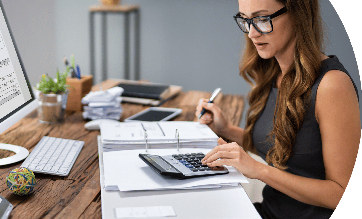 A person sitting at a desk using a calculator whilst going through some documents . In the background their computer is shown and the rest of their working environment.