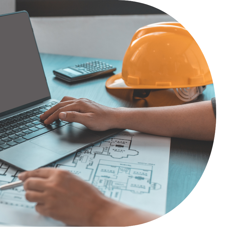 The image is cropped in a D shape and shows a close up of someones hands using a laptop, next to the laptop is a hard hat and some blue prints.