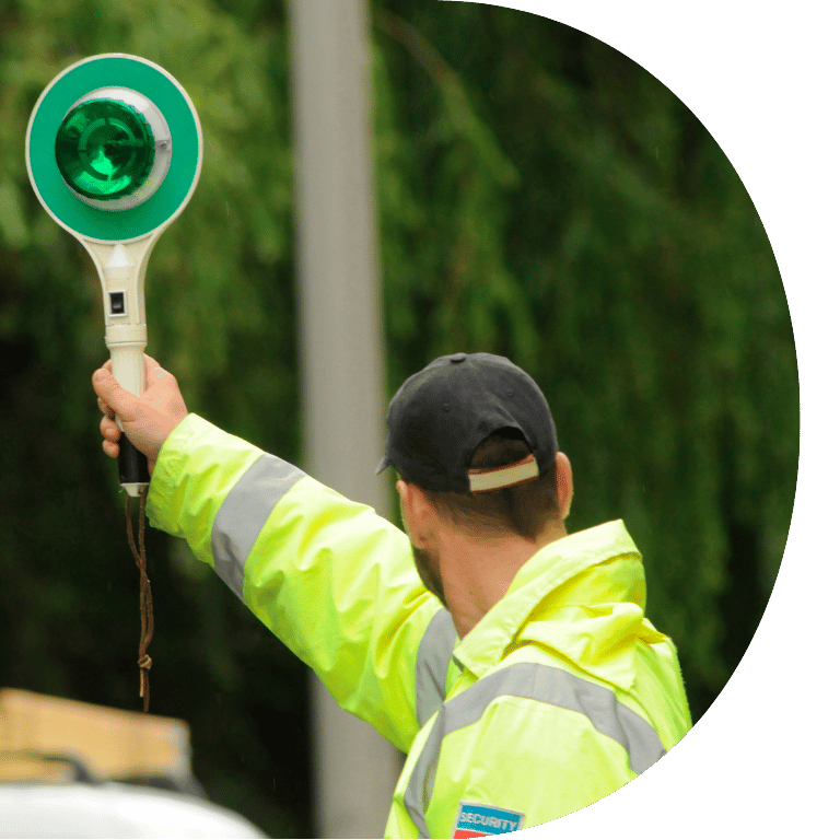 Image is cropped to a D shape and shows the back of a man in a hi--vis jacket holding a green light above his head.