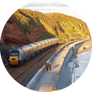 Image shows a train travelling along a coastline on a sunny day.