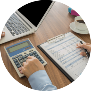 The image is cropped in a circle and shows a close up of someone using a calculator and a clipboard on a table.
