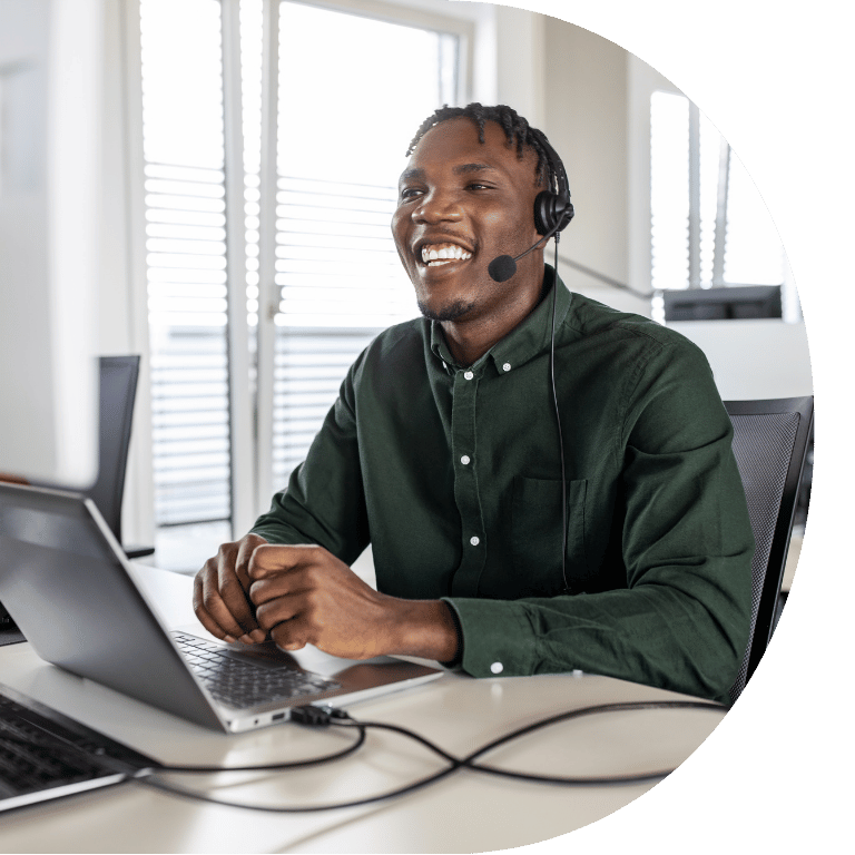 The image is cropped in a D shape and shows a person smiling behind a laptop and wearing a headset.