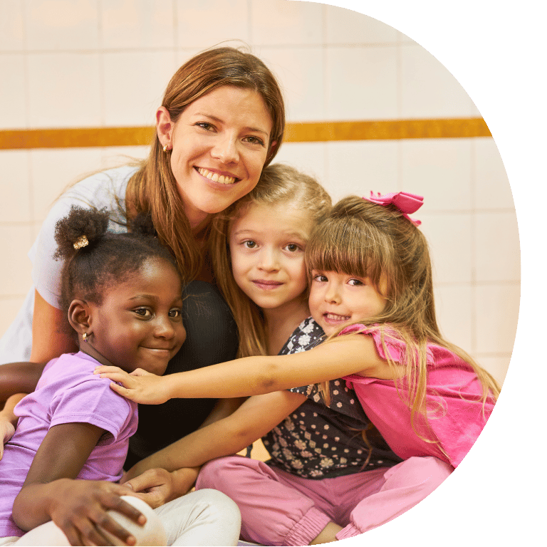 The image is cropped in a D shape and shows a teacher crouched with three students hugging.