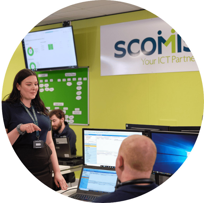 Picture of a young adult standing talking to a colleague sitting down in front of a laptop. The Scomis sign is on the wall in the background.