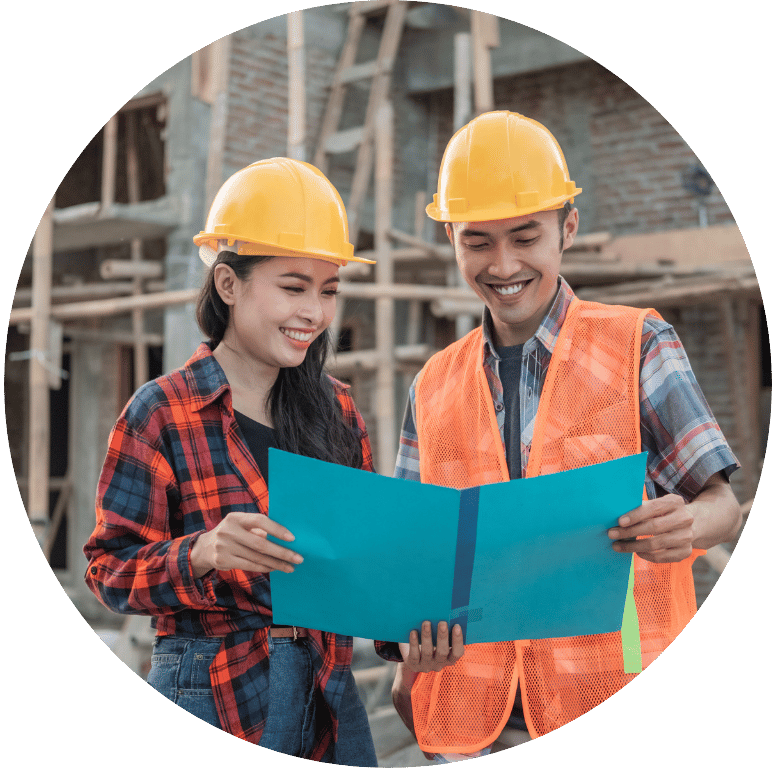 Picture of two young adults with hard hats on a construction site. They are smiling whilst looking at documents.