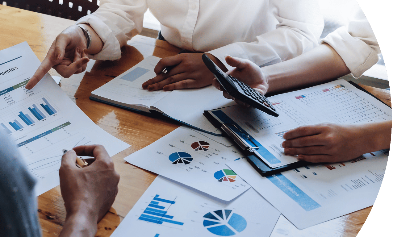 Picture of 3 people in a meeting, but only taken of their hands. There are documents on the table with graphs and information on and one is holding a calculator.