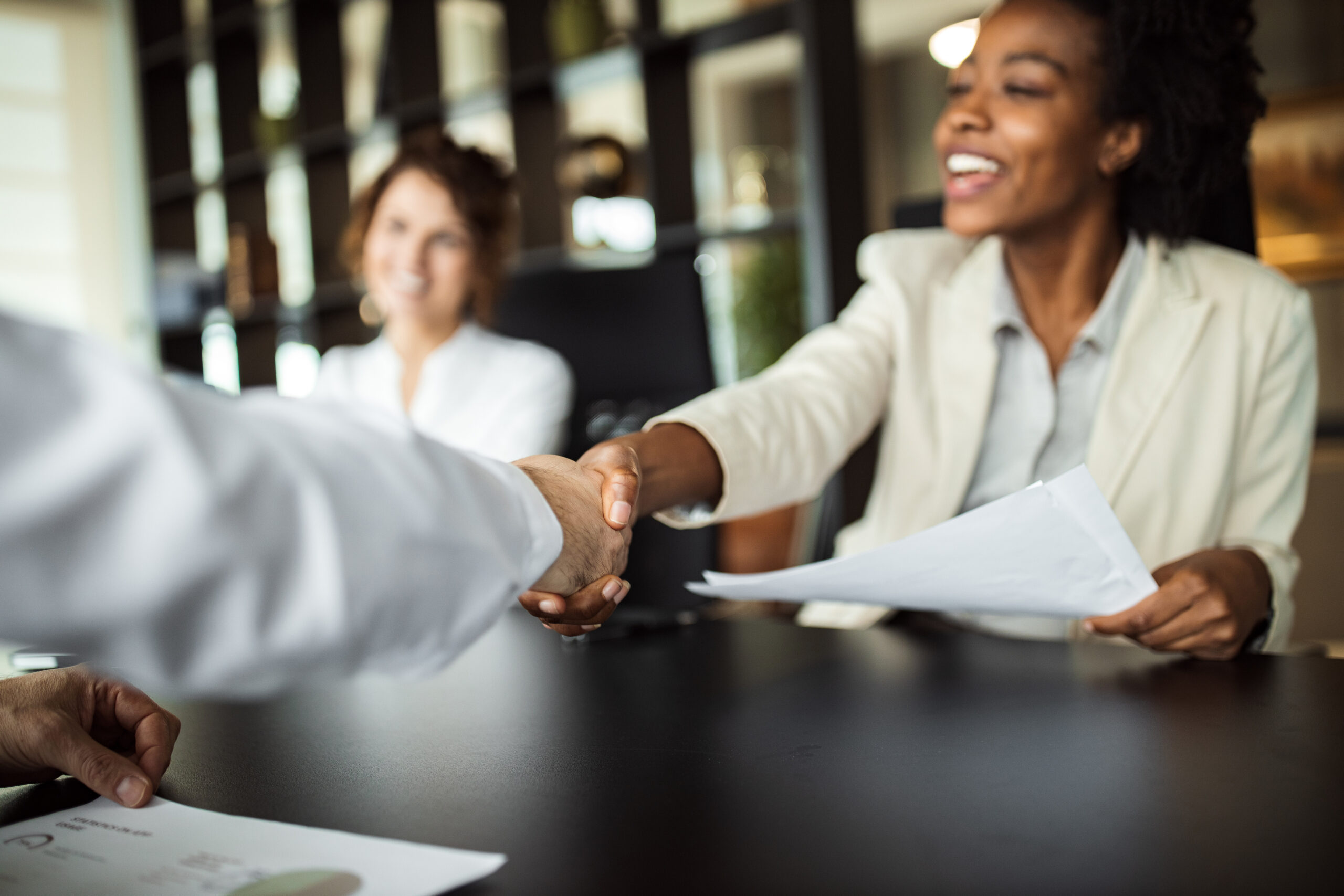 Picture of a meeting with three people in it. Two are reaching over the desk shaking hands.