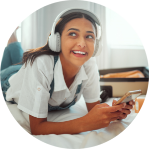 Picture of a young person lying on her stomach on the bed with headphones on and playing on her phone.