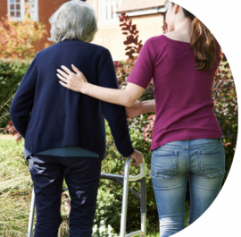 Picture of a adult helping an elderly adult using a walker. Picture taken from the back.
