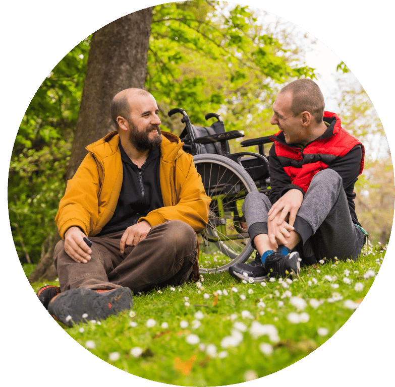 Picture of two adults sitting in a field talking to each other. In the background is a wheelchair.
