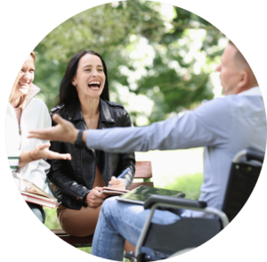 Image of two people sat on a park bench smiling and laughing with a third person in a wheelchair