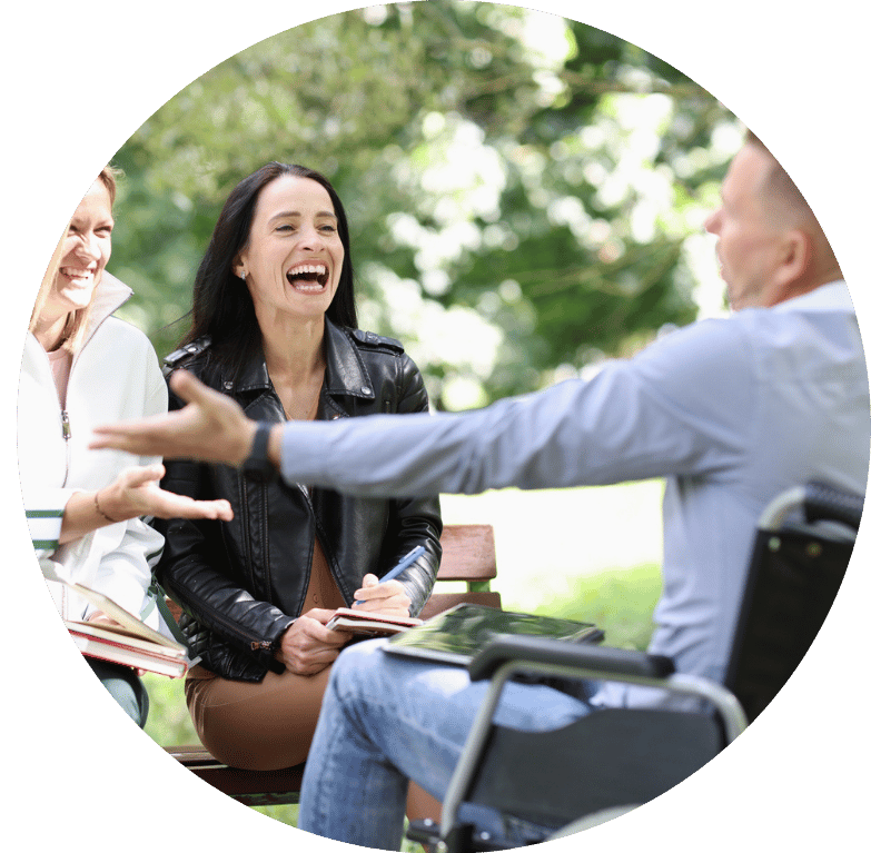 Image of two people sat on a park bench smiling and laughing with a third person in a wheelchair