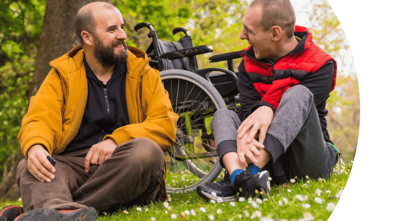 Picture of two adults sitting on grass smiling and talking to each other. One of them is a wheelchair user, which can be seen behind them.
