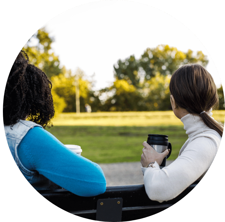 Two adults sat on a bench looking out to the fields beyond, drinking coffee together. The picture is taken from behind them, and their bodies are angled towards each other.