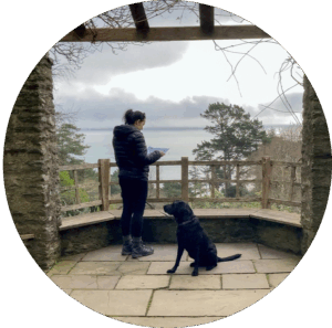 A woman reads a map on a coastal overlook on the South West Coast Path with a black labrador dog accompanying her, enjoying the serene view - Kingswear, Devon.